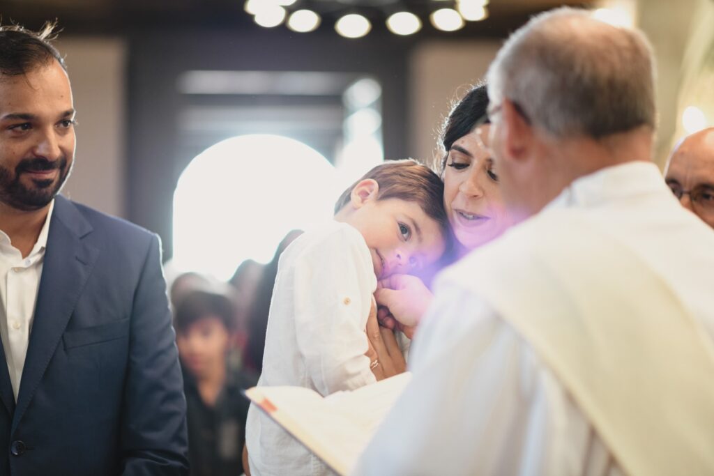 Fotografia de Batizado. Menino recebe a bênção ao colo da mãe.