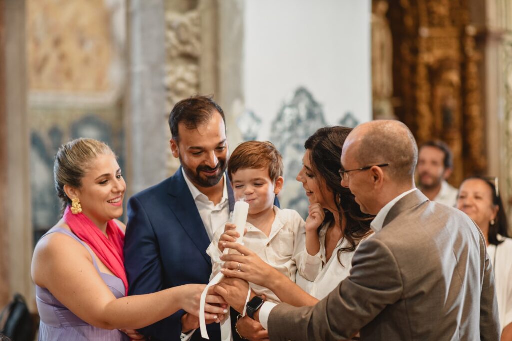 Fotografia de Batizado. Cerimónia na Igreja. Pais e padrinhos seguram a vela. Menino sopra a vela.