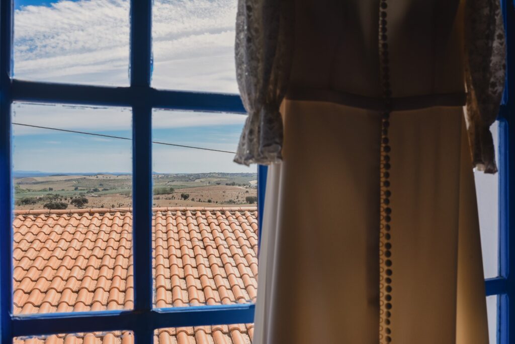 Fotografia de Casamento. Vestido da noiva pendurado à janela. Ao fundo o Alentejo.
