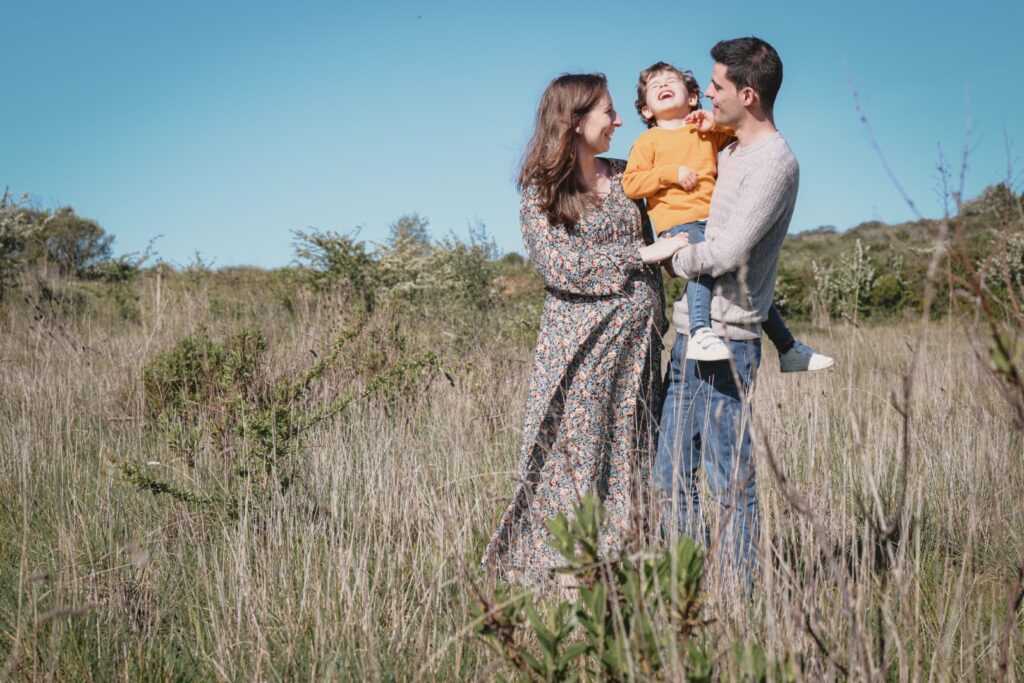 Serviços de Fotografia de Família. Sessões fotográficas em família. Sessão fotográfica em exterior. Casal espera segundo filho, com o irmão mais velho ao colo. Todos sorriem.