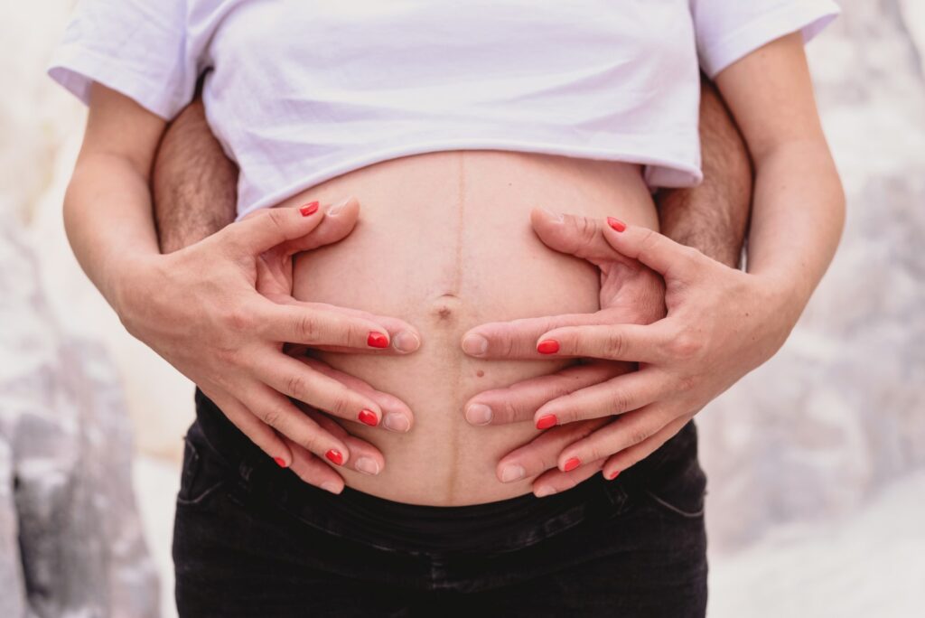 Sessões fotográficas gravidez. Sessões fotográficas em exterior de casal na natureza. Pormenor da barriga com mãos de pai e mãe.
