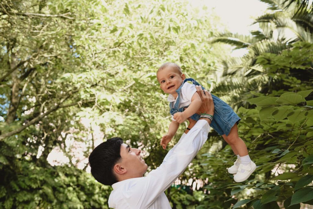 Sessões fotográficas de família. Irmão mais velho que pega o bebé ao colo. Ambos sorriem. Sessões fotográficas em exterior, num jardim.