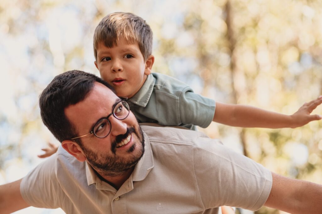 Sessões fotográficas de família. Sessões fotográficas em exterior, na natureza. 
Pai faz avião com filho às costas.